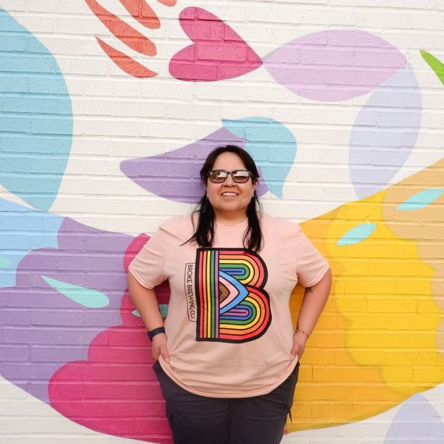 Diana De Santiago wearing a pride rainbow broke brewing co logo t-shirt posing in front of colorful mural wall